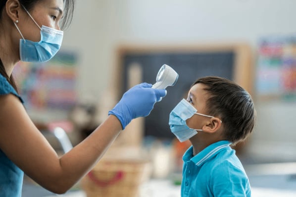 Childcare worker with mask and gloves following a daycare sick policy and taking a child's forehead temperature in the classroom.