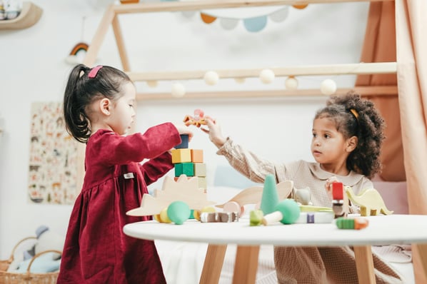 Two toddlers playing with wooden blocks at a table in a preschool classroom.