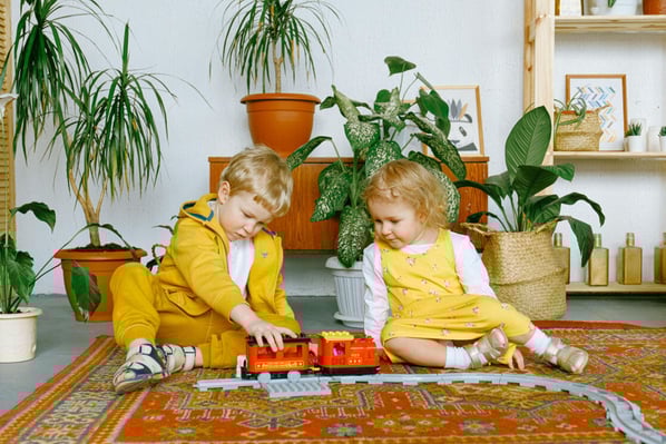 Young boy and girl dressed in bright yellow play on the floor with a toy train and tracks in a room with a lot of plants.