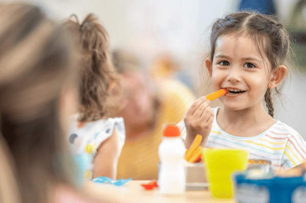 Children at daycare sitting around a table eating lunch. They have healthy eating choices on the table in front of them.