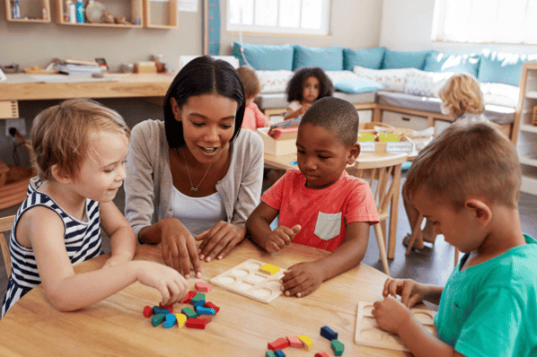 Teacher sits at a table with three children, helping them put colorful shapes into a wooden puzzle.